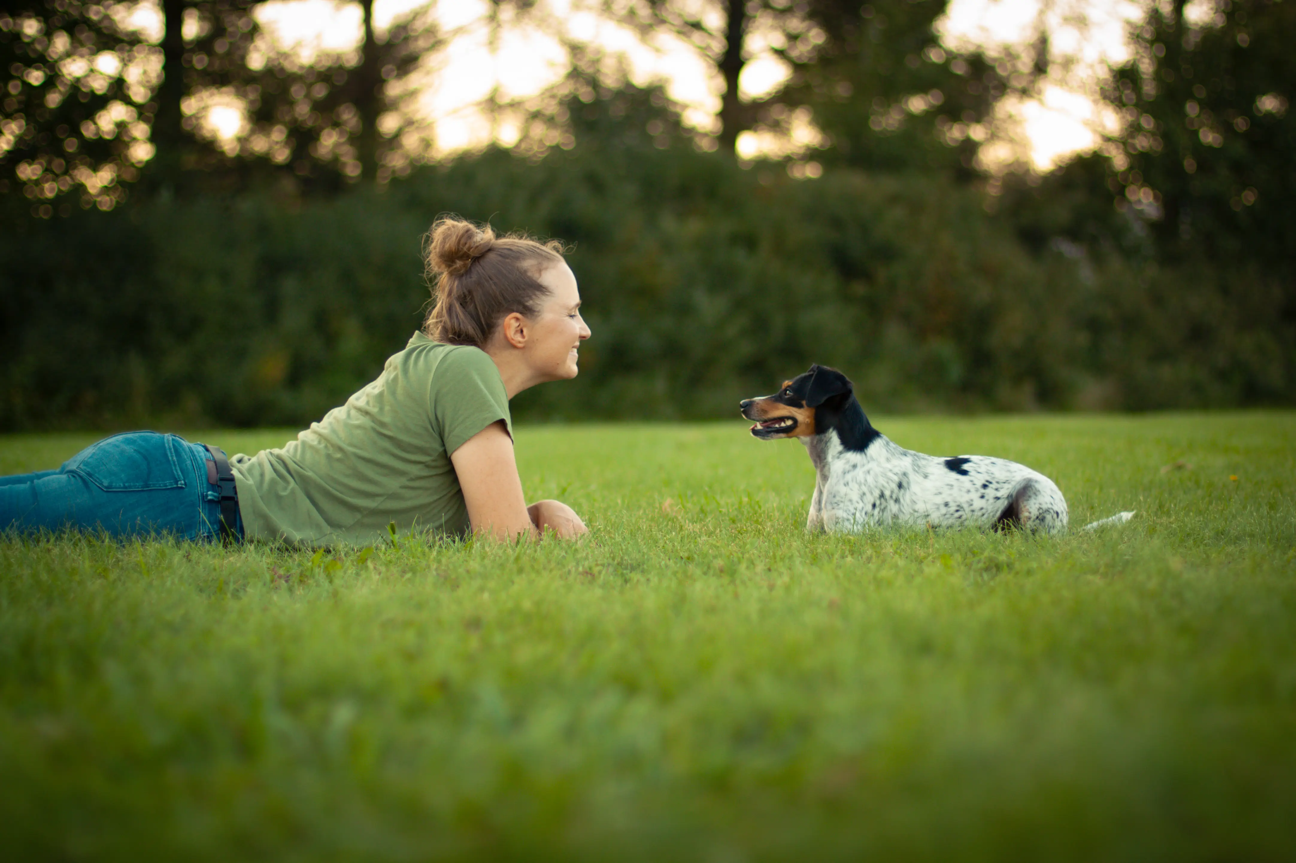 Woman with her hair in a bun, wearing a green T-shirt and jeans, lying on her stomach in a grassy field and smiling at a small black, white and tan spotted dog lying a few feet away and looking back at her.