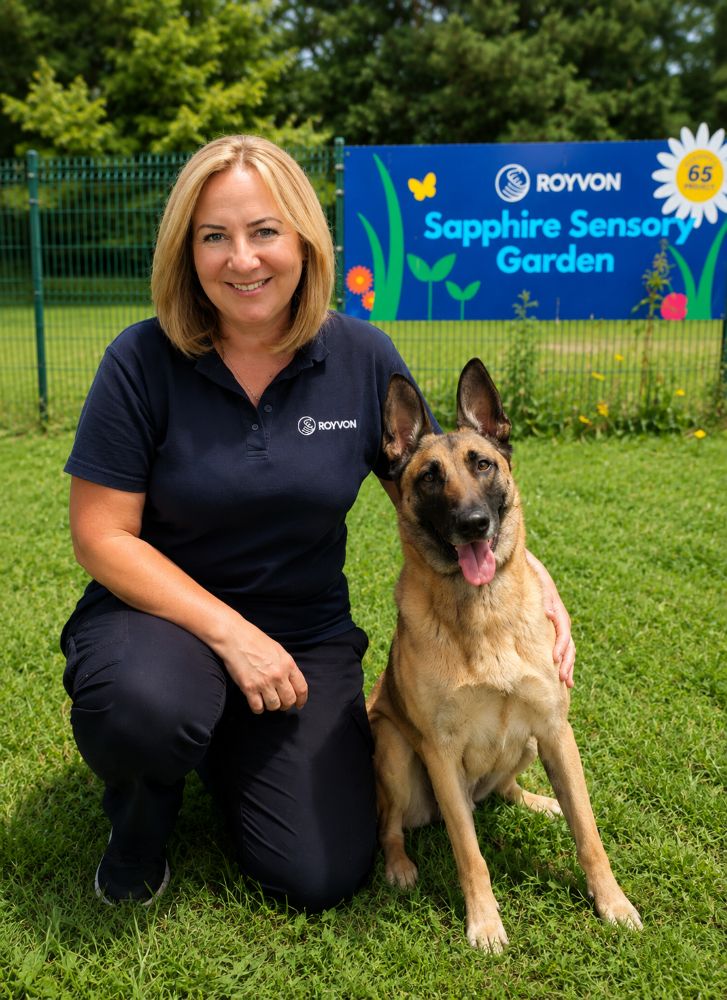 Andrea Wilde kneels on grass next to a German Shepherd in front of a blue sign that reads "Sapphire Sensory Garden." Both are smiling; the woman, who is a dog trainer, wears a navy shirt with a Royvon logo.
