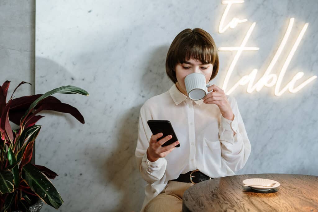 Woman enjoying coffee while using her smartphone in a cafe with neon decor and a marble wall.