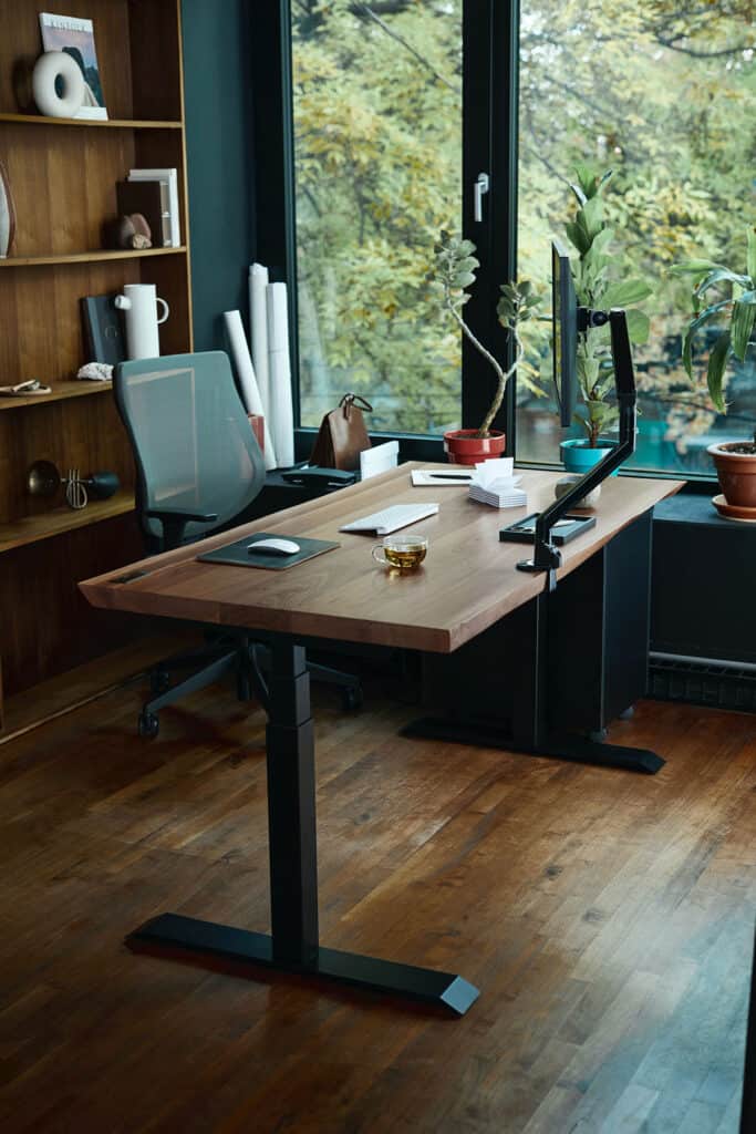 Home office with a large wooden desk, ergonomic chair, and a view of nature through large windows.