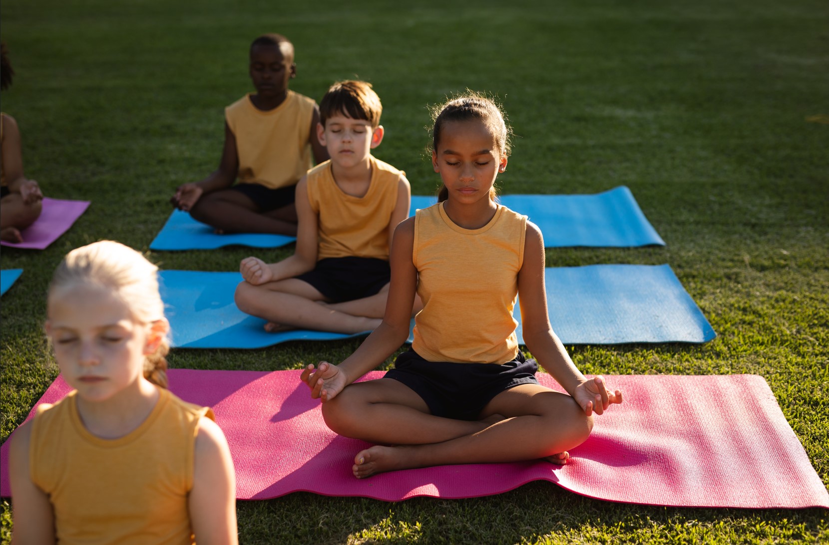 A group of children sited cross legged on yoga mats on the grass with closed eyes feeling relaxed&nbsp;